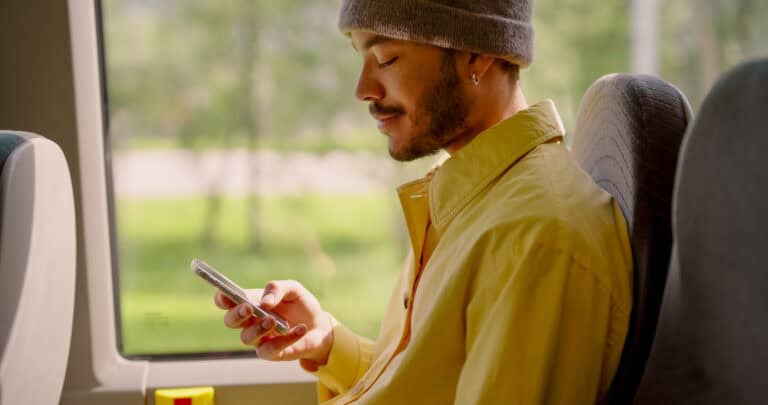 En mann iført gul skjorte og grå lue sitter ved et vindu på en buss eller et tog, ser på smarttelefonen sin og smiler litt. Grøntområder er synlige utenfor vinduet.