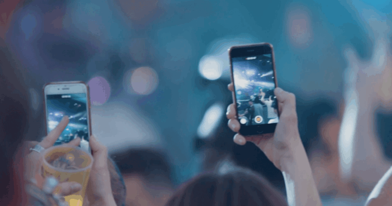 To personer på en konsert filmer scenen med smarttelefonene sine; den ene holder en drink mens han spiller inn. Den uskarpe bakgrunnen viser fargerike lys og andre publikummere.