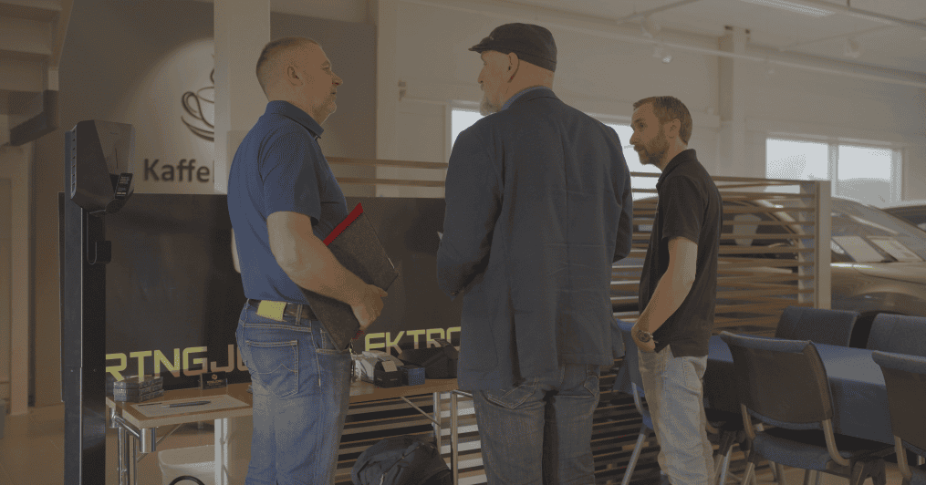 Tre menn står og snakker inne i et moderne kontor eller utstillingslokale. En mann holder et nettbrett og en mappe; en annen har på seg en caps og blazer. Stoler, bord og en skillevegg er synlige i bakgrunnen.