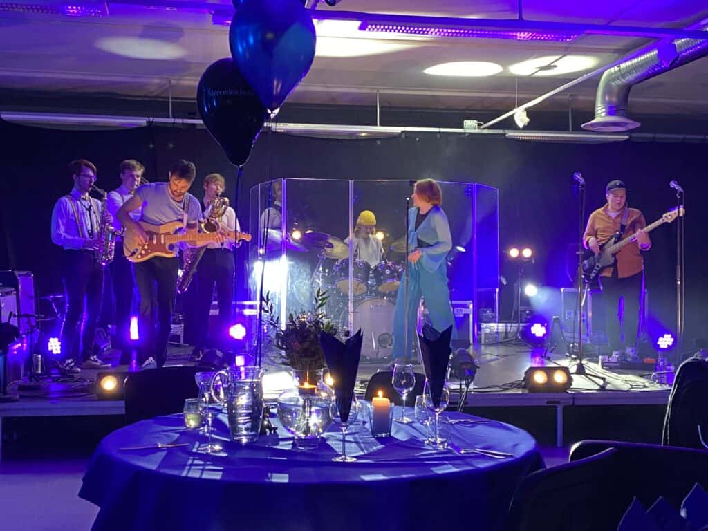 Et band opptrer på en scene med blå og lilla belysning. Musikere spiller gitar, saksofon, bass og trommer. I forgrunnen er et bord dekket med glass, stearinlys og bestikk. Ballonger svever over bordet.