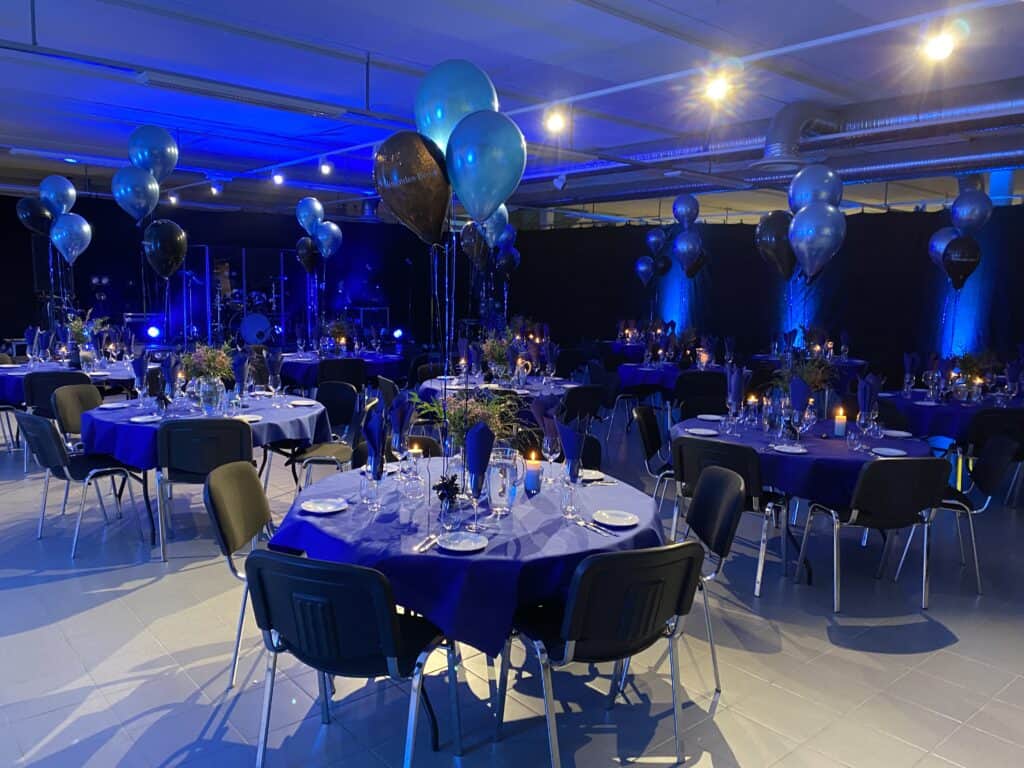 En bankettsal dekorert med blå belysning, runde bord dekket med blå duker, svarte stoler, blomsterdekorasjoner og blå eller svarte ballonger. Bordene er dekket for en formell begivenhet, med glass og lys.