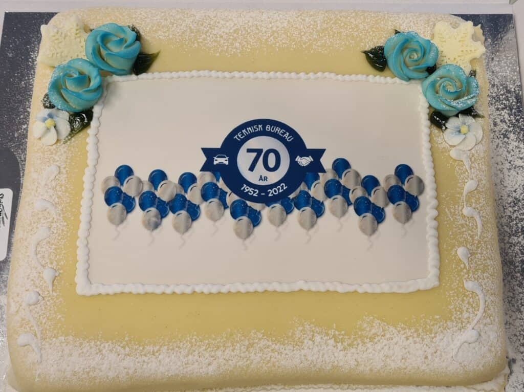 En rektangulær kake dekorert med blå og hvite roser og et trykt design med teksten Teknisk Bureau 70 År 1952-2022 i midten, omgitt av blå og hvite ballonger.