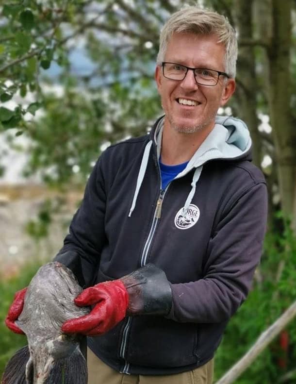 En smilende mann med briller, mørk jakke og røde hansker holder en stor fisk utendørs i nærheten av grønne trær.