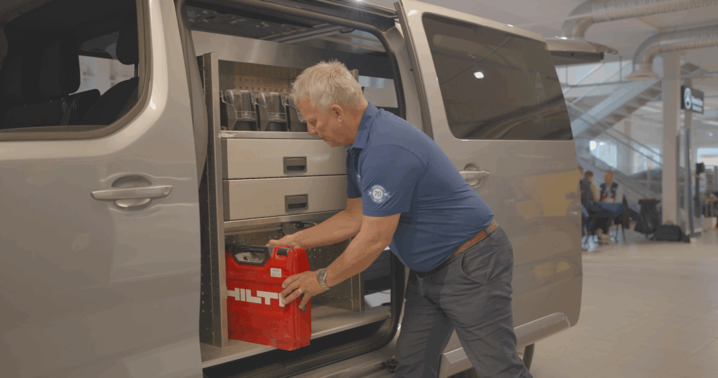 En eldre mann i blå skjorte laster en rød Hilti-verktøykasse inn i et oppbevaringsrom inne i en grå varebil med åpne skyvedører, parkert innendørs.