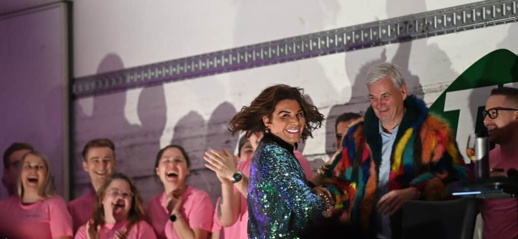 Erlend Elias i paljettjakke og en mann i fargerik pelsfrakk smiler på scenen, omgitt av en gruppe mennesker i matchende rosa skjorter som klapper og jubler.