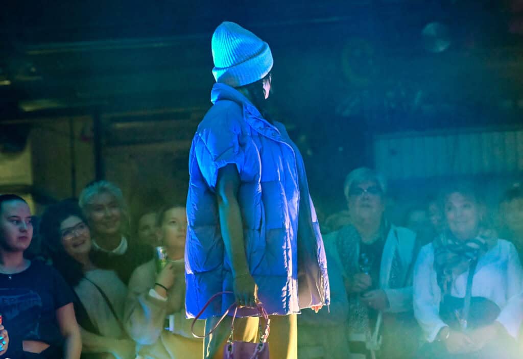 En person iført blå lue, blå dynevest og gule bukser står på scenen med en rød veske, vendt mot et publikum. Publikum ser på med forskjellige uttrykk under blå belysning.