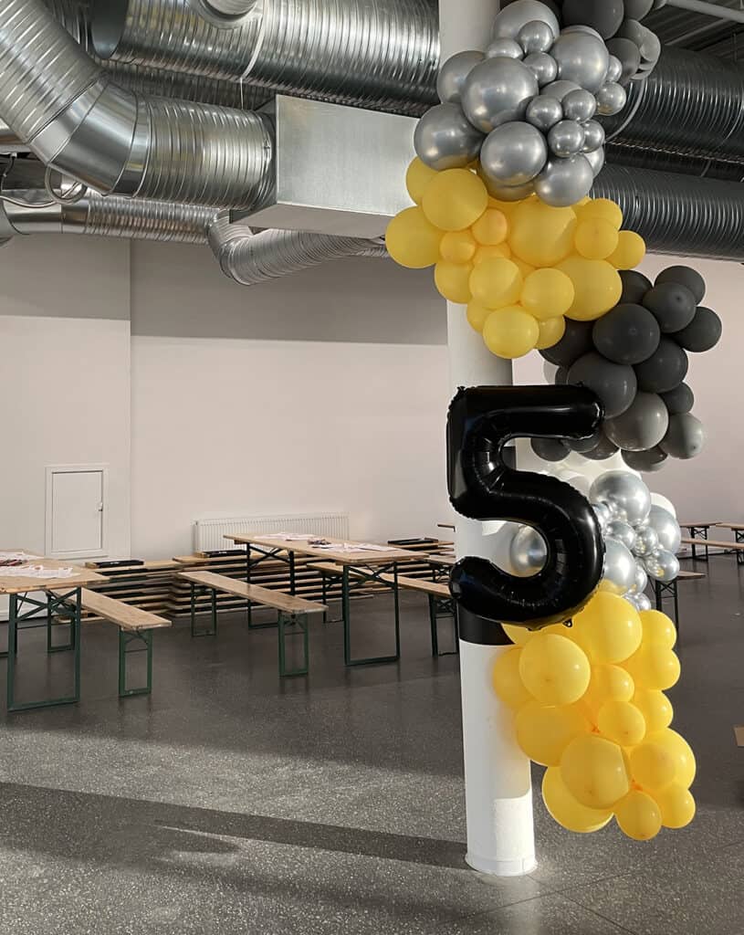 En hvit søyle dekorert med klynger av gule, svarte og sølvfargede ballonger, inkludert en stor svart ballong formet som tallet 5. I bakgrunnen er det tomme trebord og ventilasjonskanaler i metall i taket.