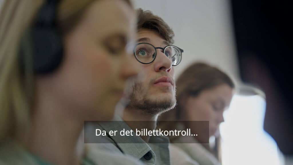 En ung mann med briller ser oppmerksomt opp mens han sitter blant andre mennesker. Den norske underteksten på bildet lyder: «Da er det billettkontroll...».