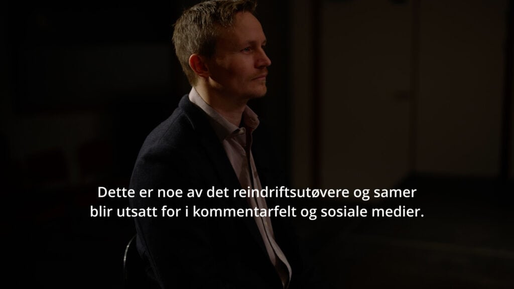 Scene fra reklamefilm i holdningskampanje mot hets og hatytringer på nett rettet mot reindriftsutøvere og samer. En mann i dress holder et ark, stående i et svakt opplyst rom med uskarpe skikkelser i bakgrunnen. Tekst sier: Dette er noe av det reindriftsutøvere og samer blir utsatt for i kommentarfelt og sosiale medier.