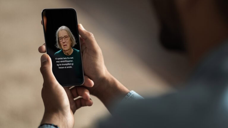 En person holder en smarttelefon som viser en video av en eldre kvinne med briller som snakker foran en mørk bakgrunn.