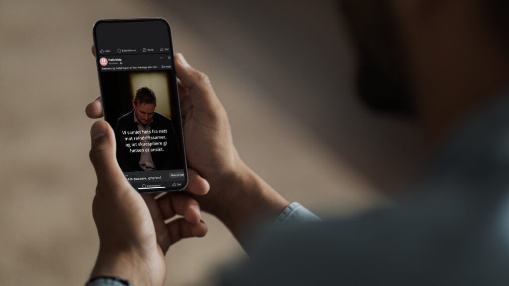 En person holder en smarttelefon som viser et bilde av en mann som leser. Scenen sees over personens skulder i svakt opplyste omgivelser.