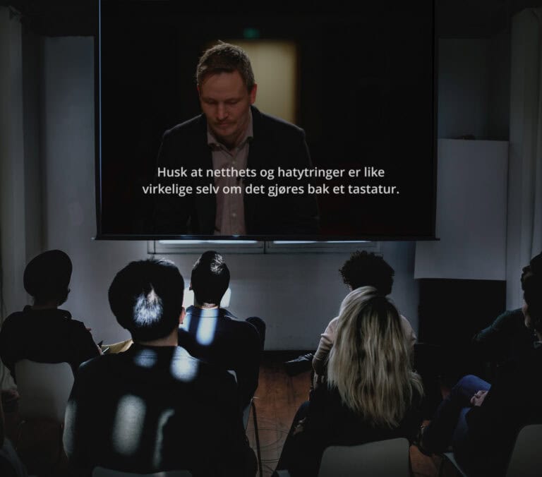 En gruppe mennesker i et mørkt rom ser på en skjerm som viser en mann med norske undertekster: Husk at netthat og trakassering er like virkelige selv om det gjøres bak et tastatur.