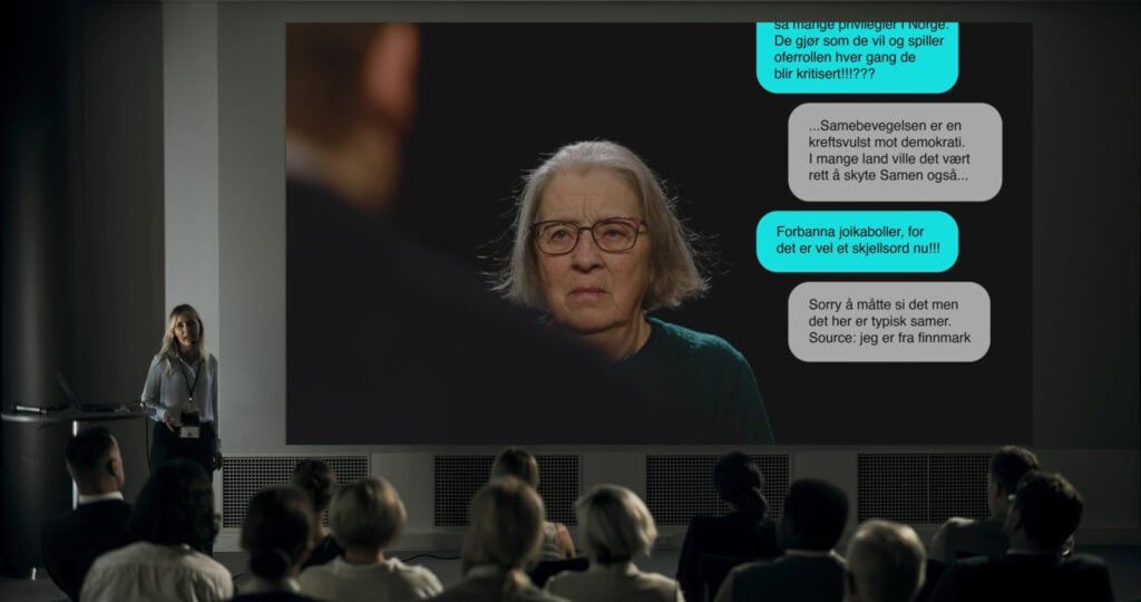 En gruppe mennesker sitter i et auditorium og ser på en presentasjon. En stor skjerm viser et nærbilde av en eldre kvinne med grått hår og briller, ved siden av tekstmeldinger.