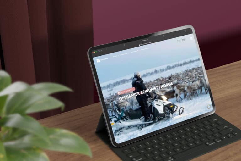 Et nettbrett med tastatur på et trebord viser nettsiden reinfakta.no med et snødekt landskap i Finnmark med en person, en snøscooter og en stor reinsdyrflokk.