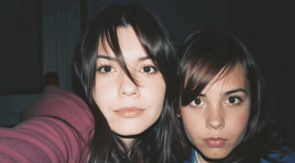 To unge kvinner med langt mørkt hår ser rett inn i kameraet og poserer tett sammen for en selfie mot en mørk bakgrunn. Den ene har på seg en rødbrun hettegenser, og den andre har pannelugg og et alvorlig uttrykk. Bildet er AI generert.