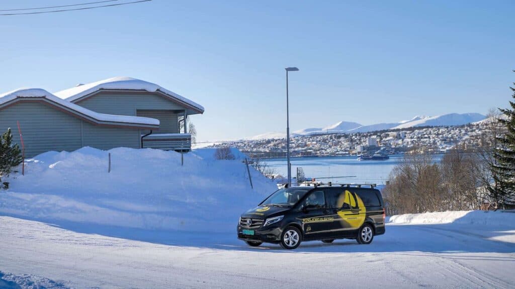 En svart varebil med gul grafikk står parkert på en snødekt vei, med snødekte hus og store snøfonner i nærheten. I bakgrunnen er det utsikt over en by og fjell under en klar blå himmel.
