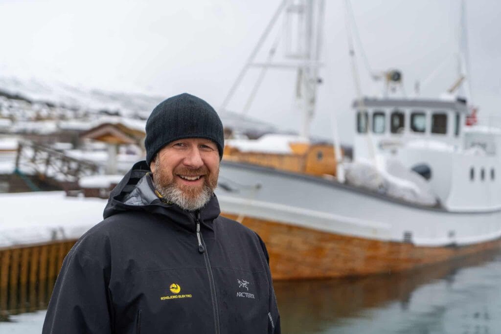 En skjeggete mann i svart lue og vinterjakke smiler mens han står foran en fiskebåt av tre som ligger fortøyd i en snødekt havn. Snødekte fjell er synlige i bakgrunnen.