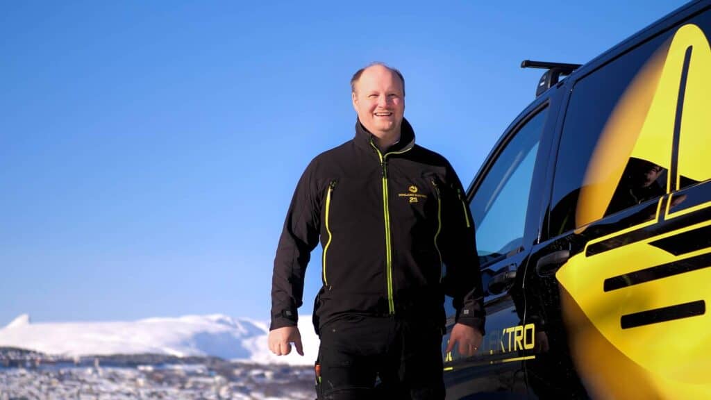 En smilende mann i svart jakke står utendørs ved siden av et svart kjøretøy med gul grafikk, med snødekte fjell og en klar blå himmel i bakgrunnen.