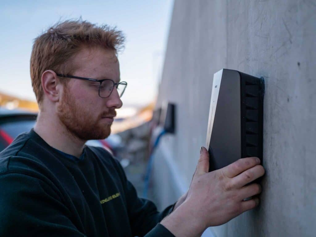 En mann med briller og lysebrunt hår installerer en rektangulær svart enhet på en betongvegg utendørs, og fokuserer intenst på arbeidet sitt.