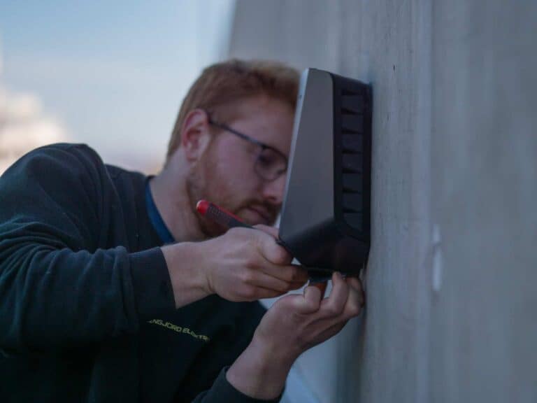 En person med briller bruker en skrutrekker til å installere eller reparere en svart enhet montert på en grå vegg, og fokuserer intenst på oppgaven.