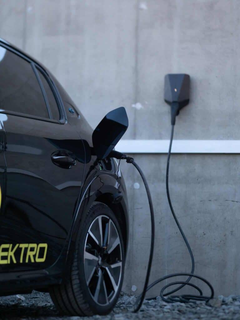 En svart elbil står parkert ved siden av en betongvegg og lades via en kabel koblet til en veggmontert ladestasjon. Bare baksiden av kjøretøyet er synlig.