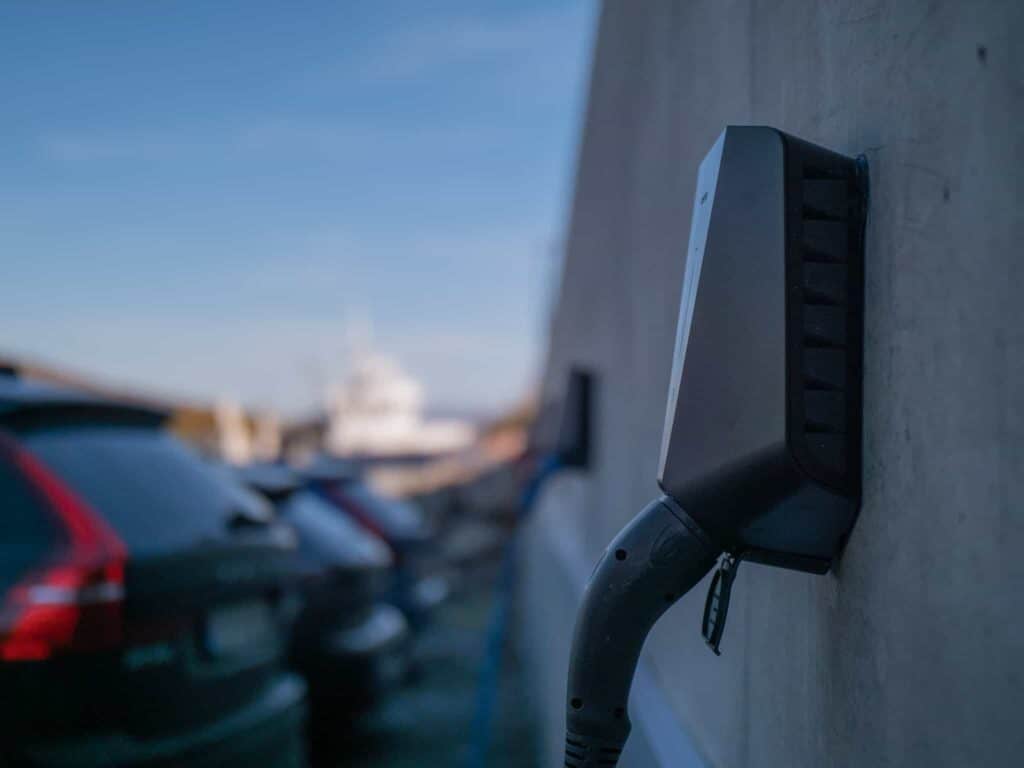 Et nærbilde av en ladestasjon for elbiler montert på en vegg, med parkerte biler og en uskarp båt i bakgrunnen på en solskinnsdag.