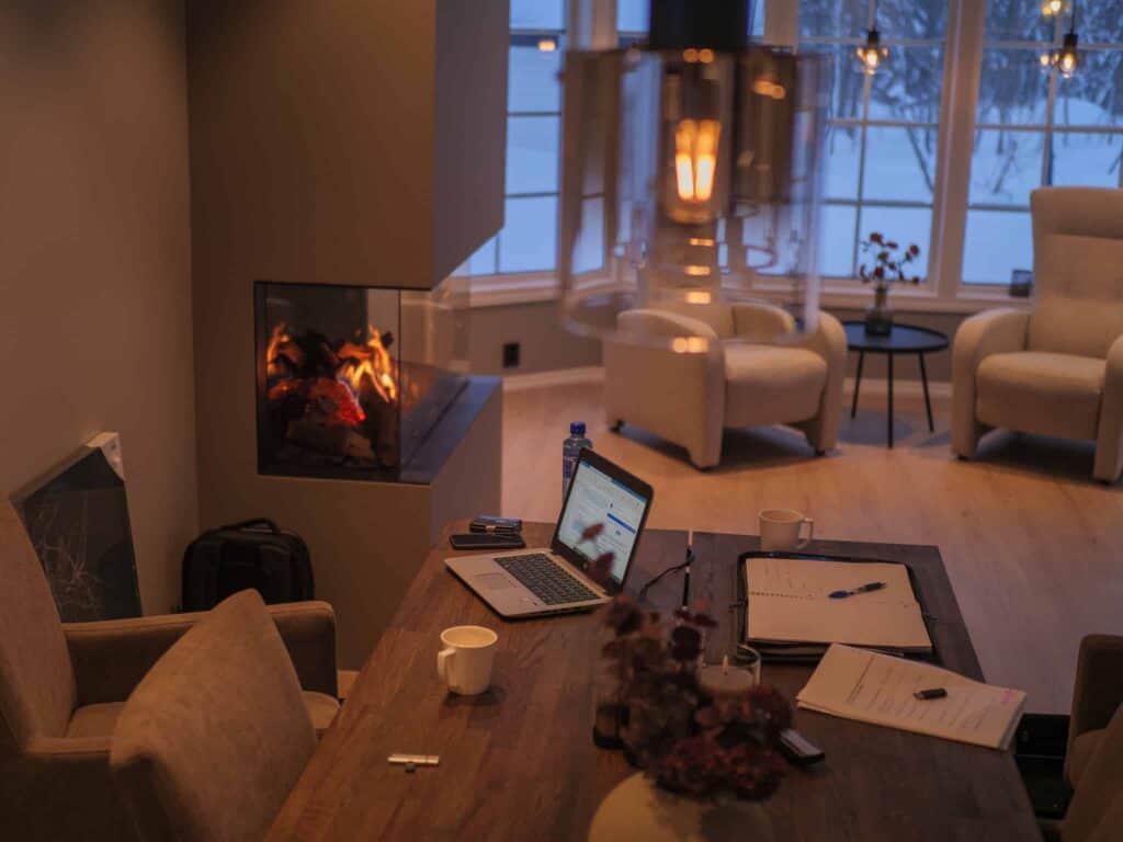 Et koselig rom med en moderne peis, et trebord med en bærbar PC, papirer, krus og en penn. Myke lenestoler står ved store vinduer og viser frem et snødekt utendørsscene. Varmt, stemningsfullt lys fyller rommet.