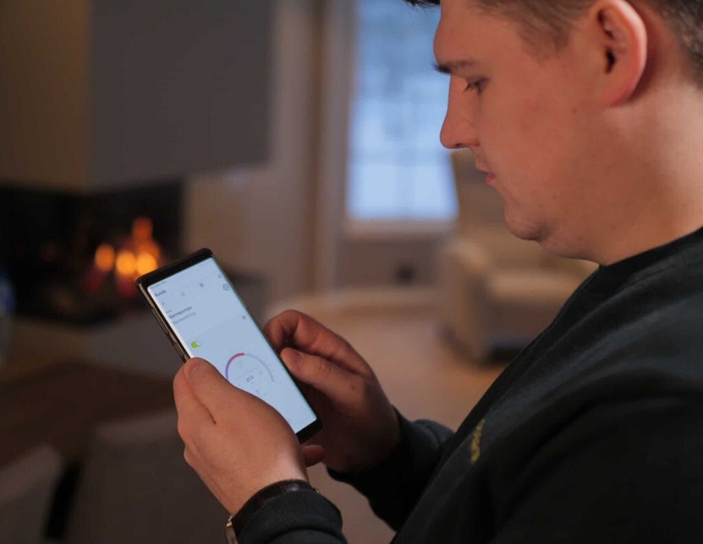 En mann står innendørs og bruker smarttelefonen sin. Han navigerer i en app med en sirkulær graf på skjermen. Bakgrunnen viser en mykt opplyst stue med peis.