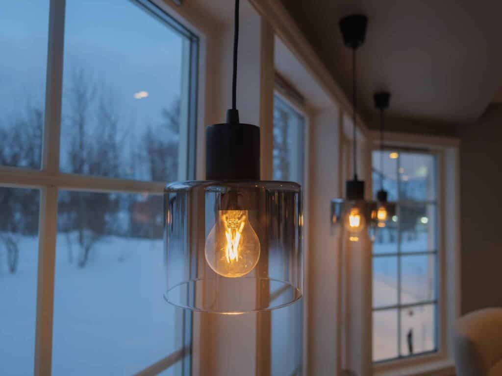 Et nærbilde av en hengende pendellampe med en synlig pære som lyser varmt innendørs nær store vinduer. Snø er synlig utenfor, og to pendellamper til henger i bakgrunnen.