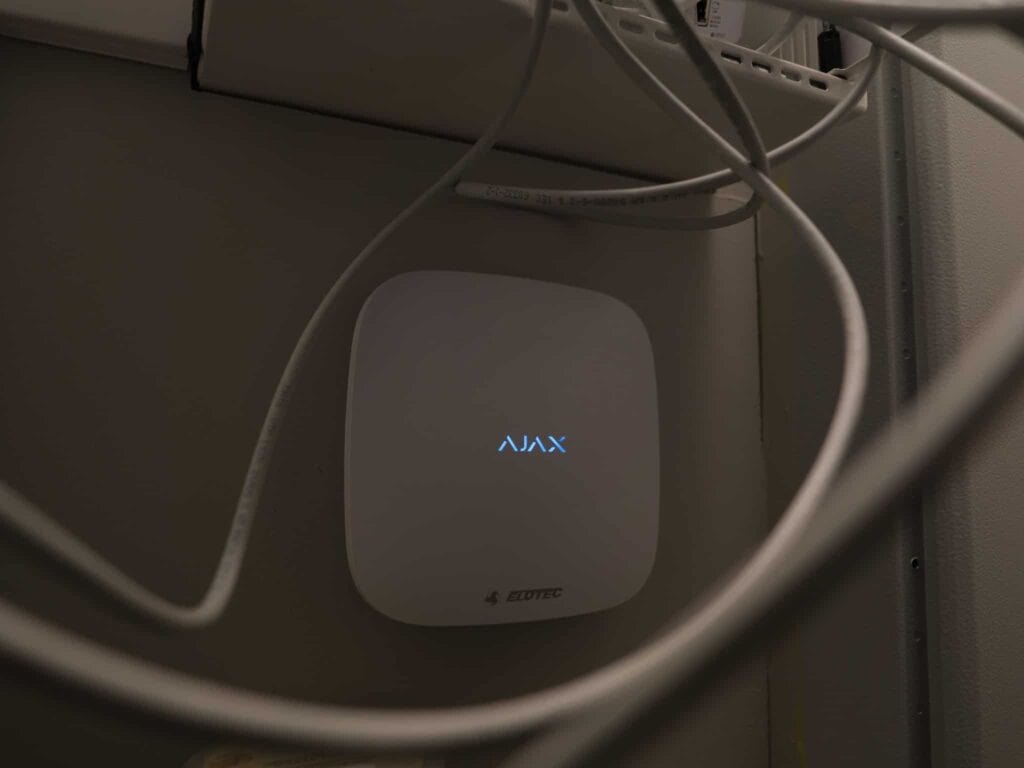 Et nærbilde av en hvit elektronisk enhet merket AJAX med et lite blått lys, montert på en vegg blant flere nettverkskabler. Området rundt ser ut til å være et verktøy- eller serverskap.