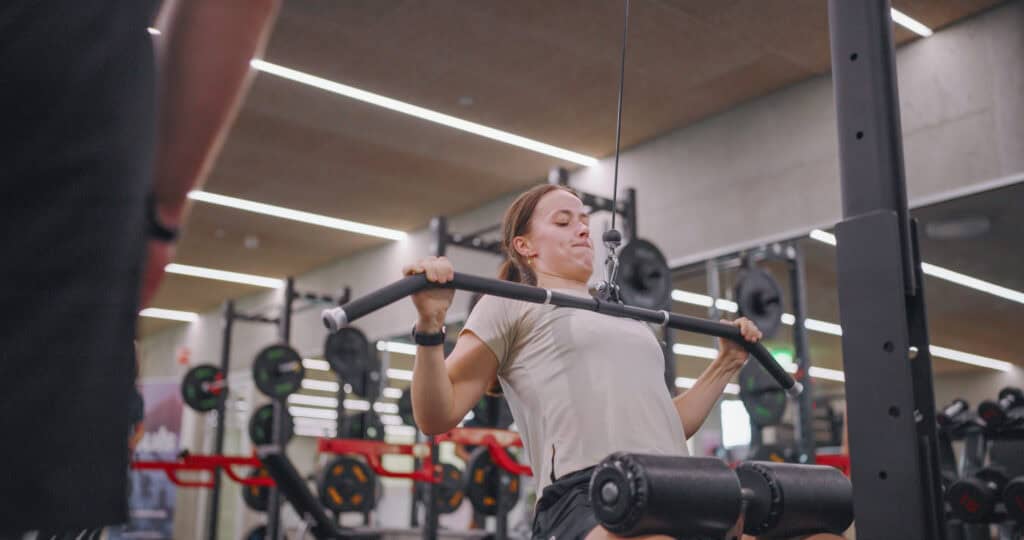 En kvinne i treningstøy trener på en lat pulldown-maskin i et treningsstudio og trekker stangen ned med begge hender. Vektskiver og treningsutstyr er synlige i bakgrunnen.