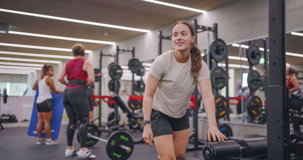 En kvinne i en lys skjorte og svarte shorts smiler mens hun hviler hånden på treningsutstyr i et treningssenter. To andre kvinner trener i bakgrunnen i nærheten av vektløfterstativer.