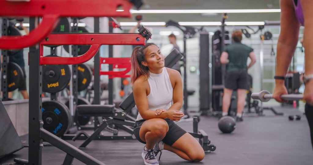 En kvinne i sportstøy sitter på huk og smiler mens hun snakker med en annen person som løfter en vektstang i et treningsstudio fylt med diverse treningsutstyr.