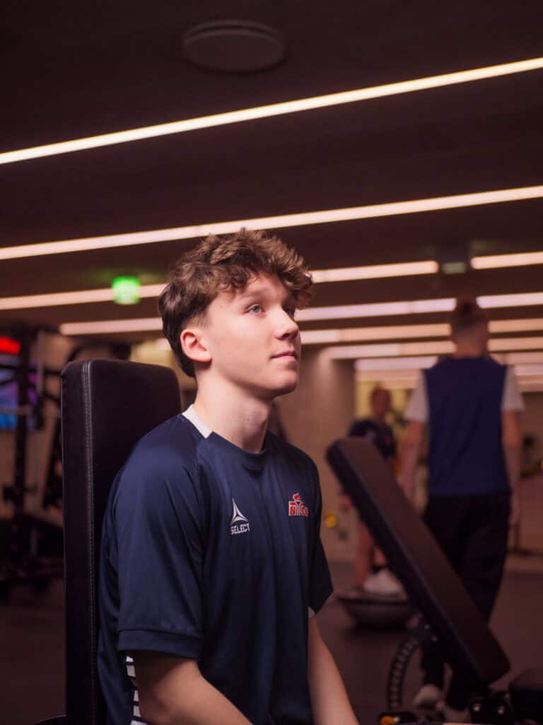 En ung mann med krøllete hår, iført en marineblå sportsskjorte, sitter på treningsutstyr og ser oppover. Treningsstudioet har sterke taklamper og andre personer som trener i bakgrunnen.