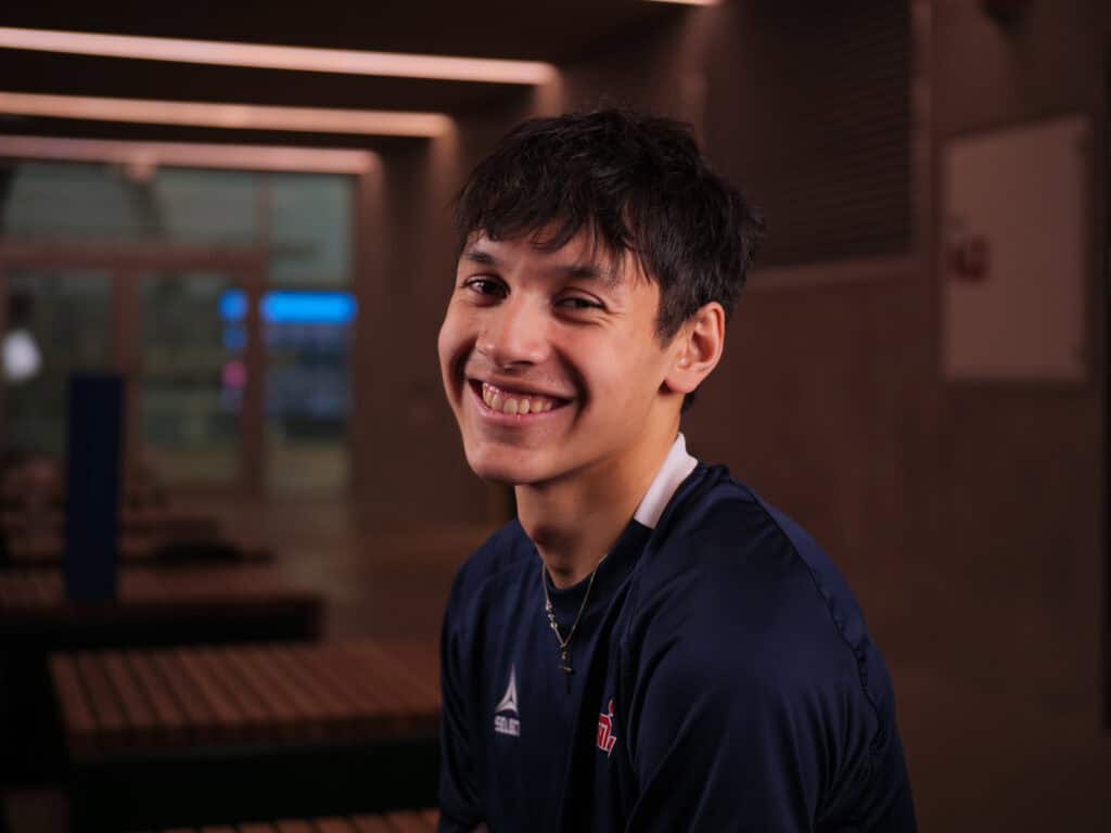En ung person med kort mørkt hår iført en marineblå atletisk skjorte smiler varmt mens han sitter innendørs på en trebenk, med mykt lys og en uskarp bakgrunn.