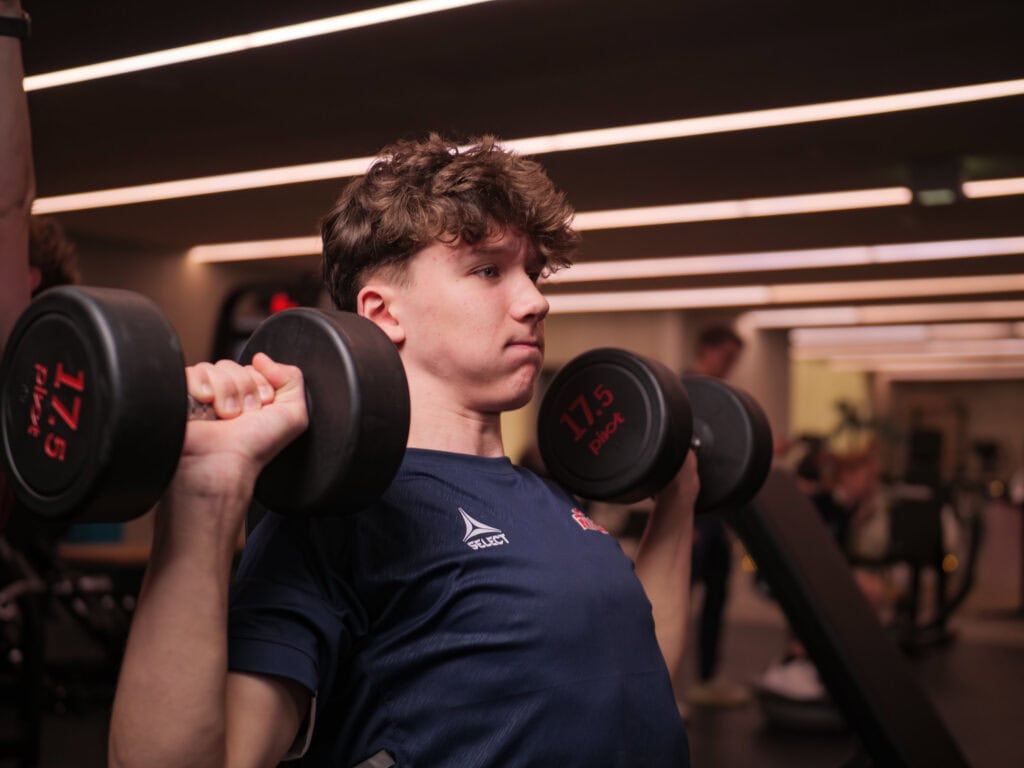 En ung mann med krøllete hår løfter to 17,5 kg tunge manualer i skulderhøyde i et treningsstudio, iført en marineblå skjorte. Bakgrunnen i treningsstudioet er mykt opplyst av lineære taklamper.