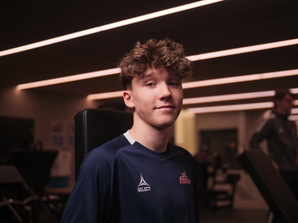 En ung person med krøllete hår og et svakt smil sitter i et treningsstudio, iført en mørk sportsskjorte. Bakgrunnen viser treningsutstyr og varme, lineære taklamper.