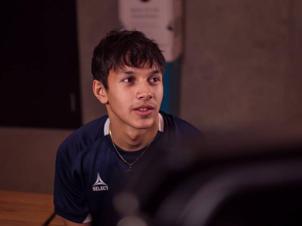 En ung mann med kort mørkt hår iført en marineblå sportstrøye sitter innendørs og ser litt oppover og til siden med et nøytralt uttrykk.