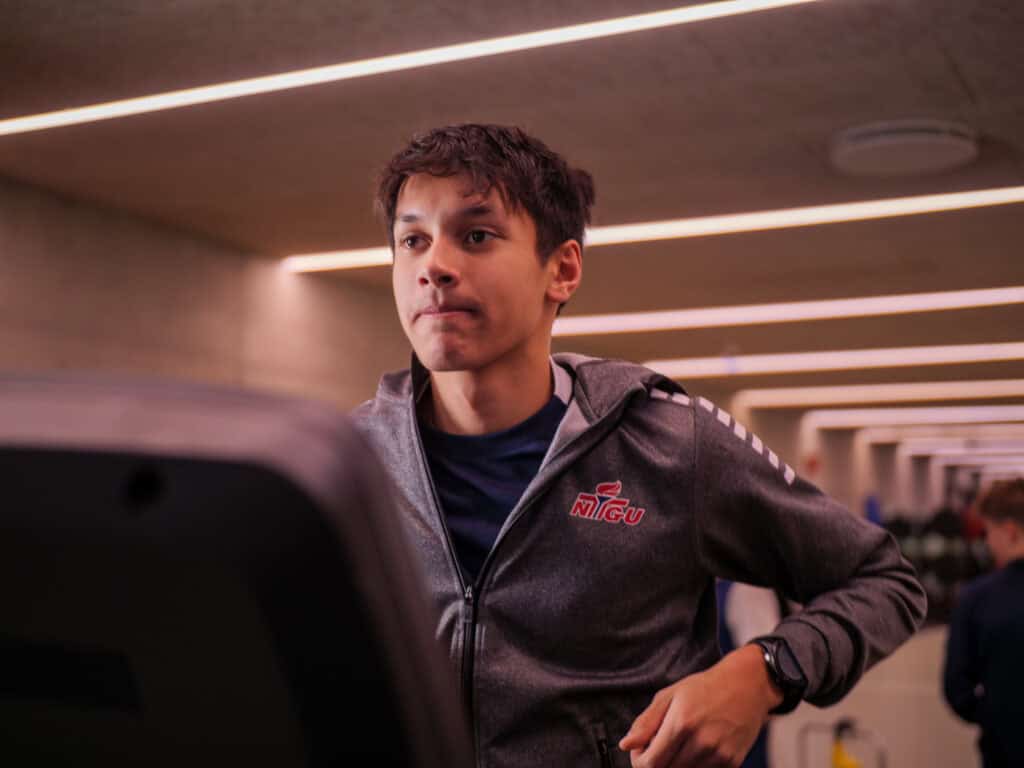 En ung mann i grå jakke med logo løper på tredemølle innendørs og ser fokusert ut. Sterke taklamper og uskarpe mennesker er synlige i bakgrunnen.