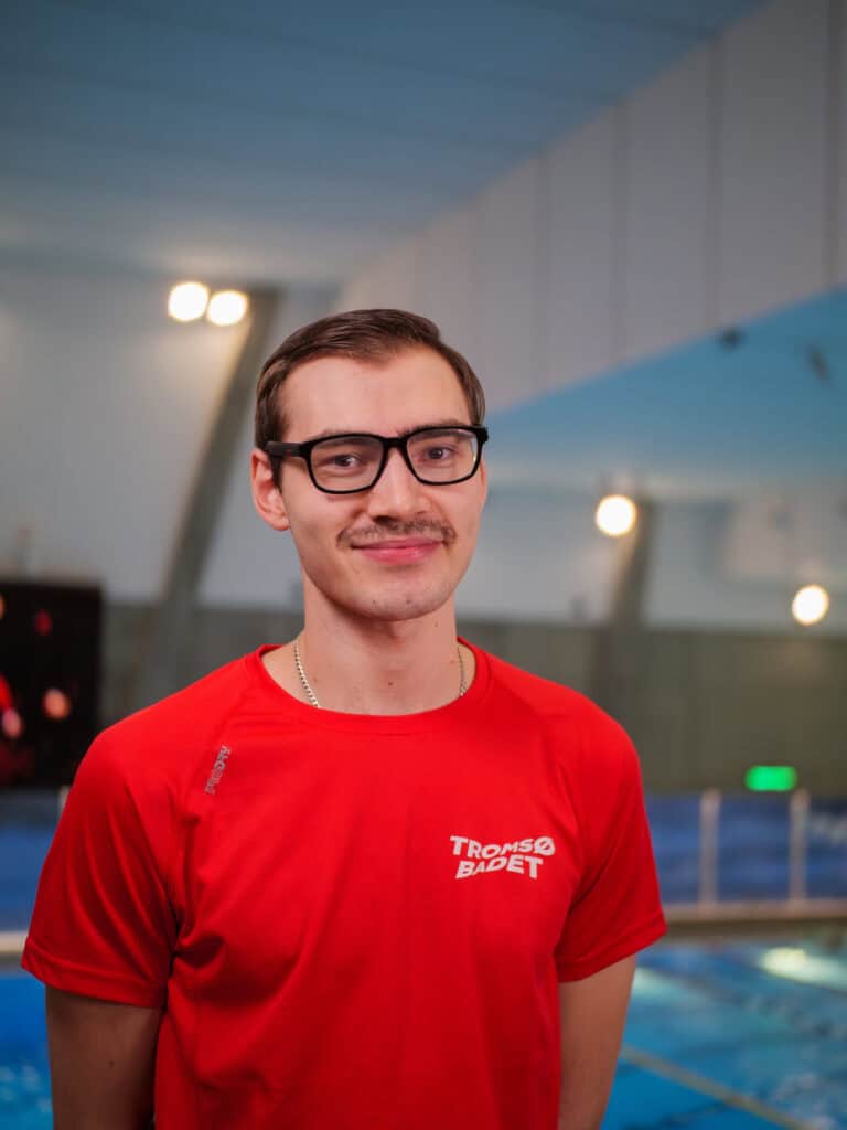En ung mann med kort brunt hår, briller og bart står innendørs ved et svømmebasseng, iført en rød Tromsø Badet-t-skjorte og smiler mykt til kameraet.