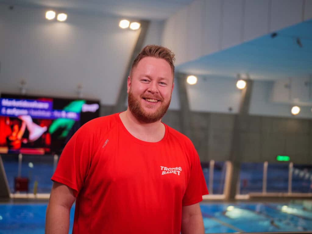 En smilende mann med skjegg, iført en rød Tromsø Badet-skjorte, står foran et innendørs svømmebasseng med blått vann og sterke lys i bakgrunnen.