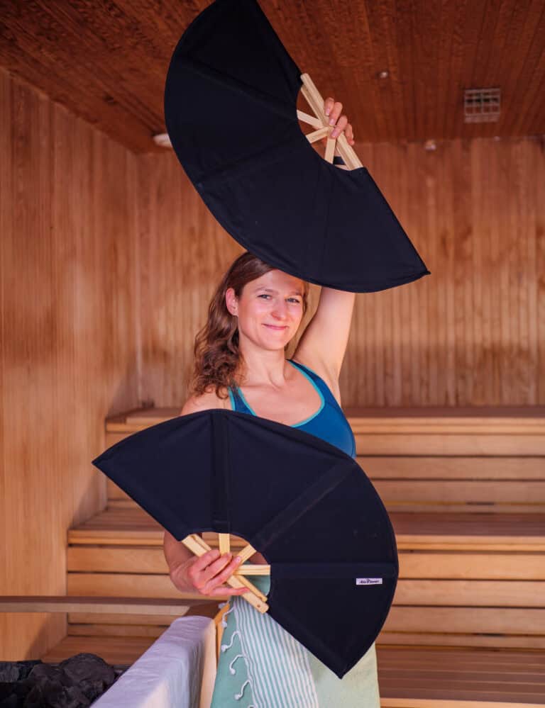 En kvinne i en badstue holder to store, svarte vifter, en over hodet og en foran brystet. Hun har på seg en blå singlet og et håndkle surret rundt livet. Badstuen har trevegger og benker.