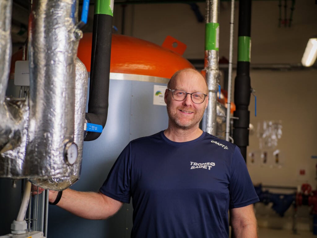 En smilende mann med briller og en blå Tromsø Badet-skjorte står i et industrielt miljø med isolerte rør og en stor oransje tank i bakgrunnen.
