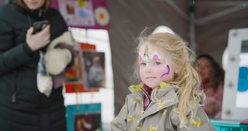 Et lite barn med blondt hår og fargerik ansiktsmaling står innendørs iført en regnfrakk. I bakgrunnen holder en voksen en telefon og et kosedyr. Fargefull kunst og dekorasjoner er synlige bak dem.