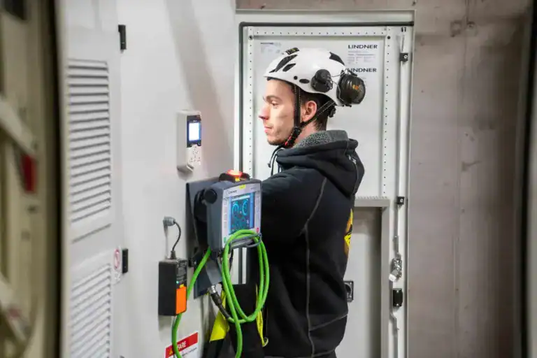 En arbeider iført sikkerhetshjelm og svart hettegenser betjener elektrisk utstyr i et industrielt miljø, stående foran et åpent kontrollpanel med diverse kabler og enheter tilkoblet.