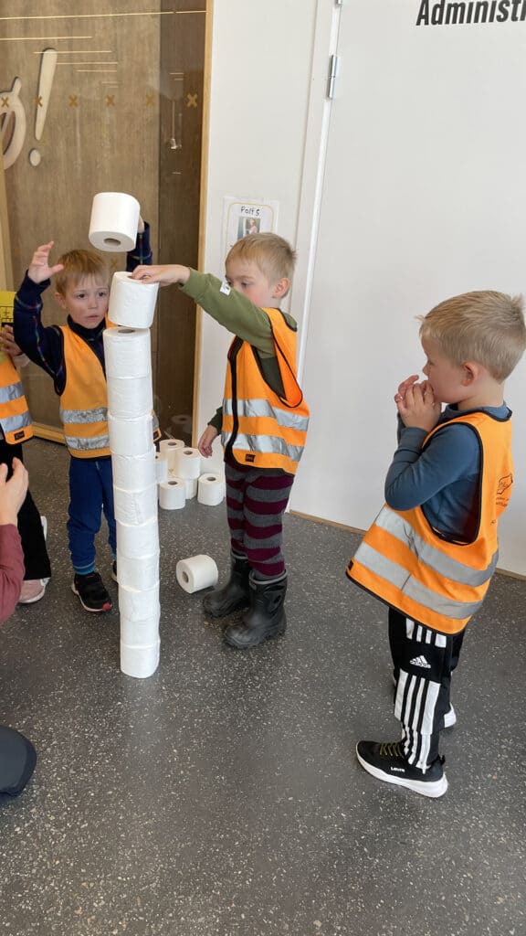 Tre små barn i oransje sikkerhetsvester stabler toalettruller inn i et høyt tårn innendørs. Ett barn legger en rull på toppen mens de andre ser på.