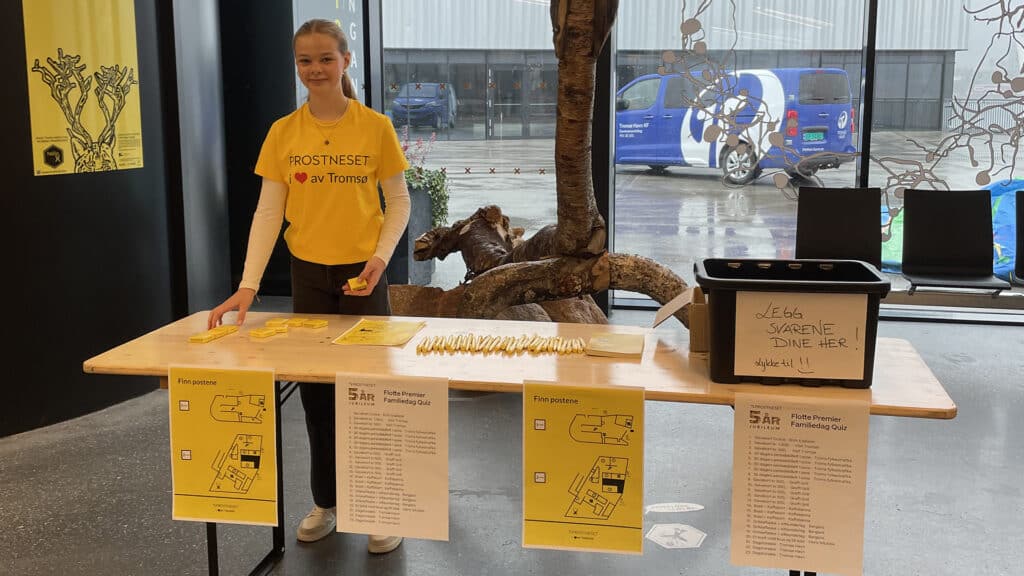 En ung kvinne i gul skjorte står bak et bord med papirer og nøkkelringer, klar til å hjelpe. Bordet har kart og en eske merket «Leg Svaret Dine Her». Scenen er innendørs, med vinduer som viser biler utenfor.