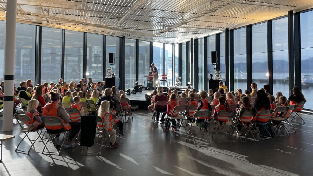 En gruppe barn og voksne, mange iført refleksvester, sitter i stoler vendt mot en taler i et stort, solfylt rom med gulv-til-tak-vinduer og en naturskjønn utsikt utendørs.