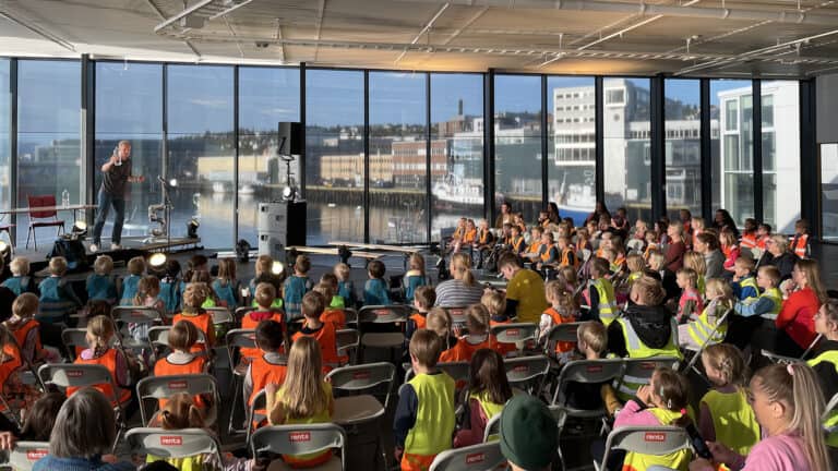 En utøver står på scenen foran store vinduer med utsikt over en havn og underholder et sittende publikum av små barn iført fargerike vester inne i et lyst, moderne rom.