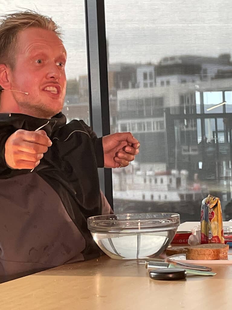 En mann med headsetmikrofon gestikulerer livlig mens han sitter ved et bord med en bolle med vann, husholdningsartikler og en tube sennep; bybygninger er synlige gjennom vinduet bak ham.